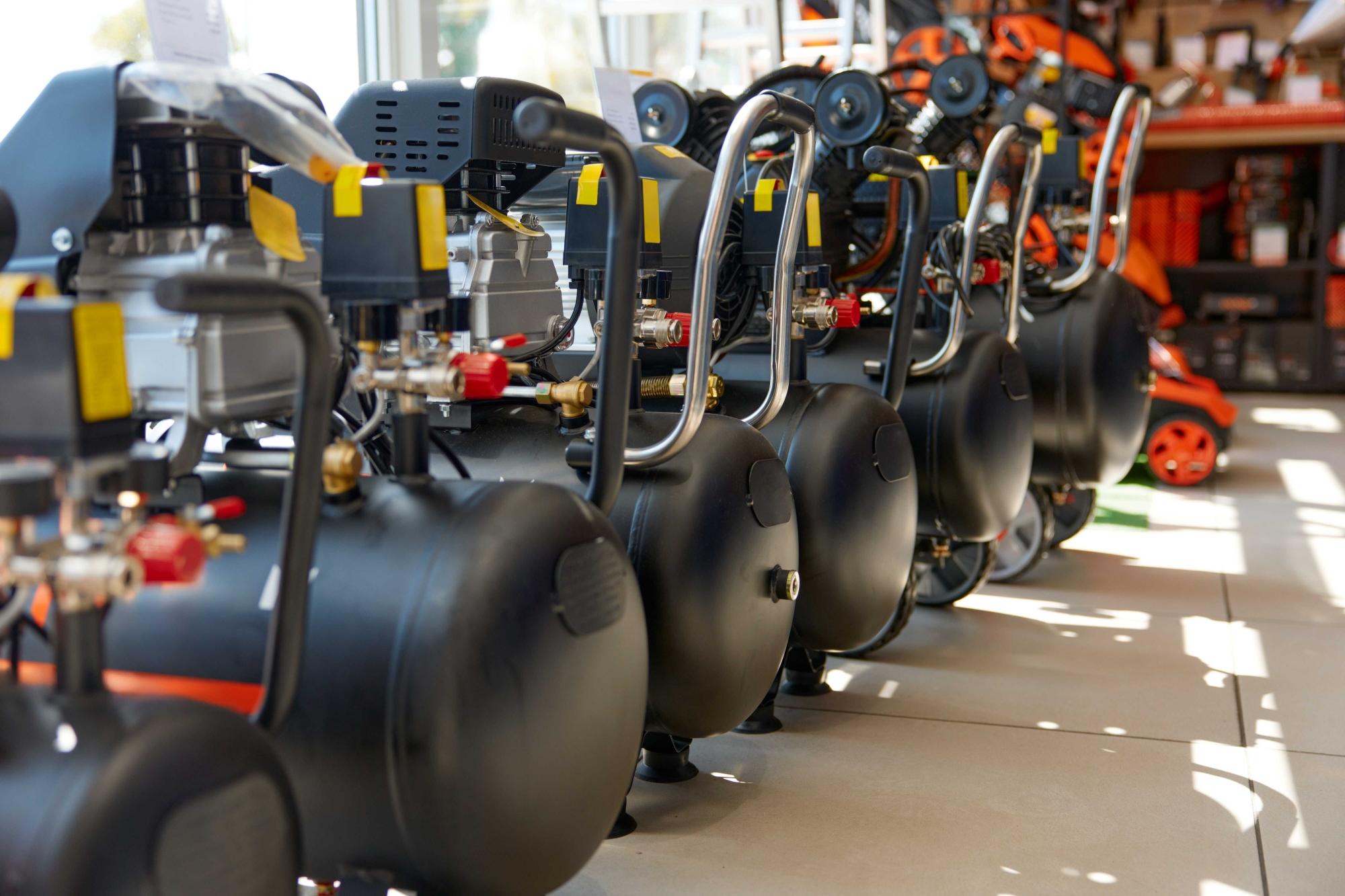 Row of large black industrial gas or air tanks with hoses, valves, and gauges arranged in a workshop showroom or service area.